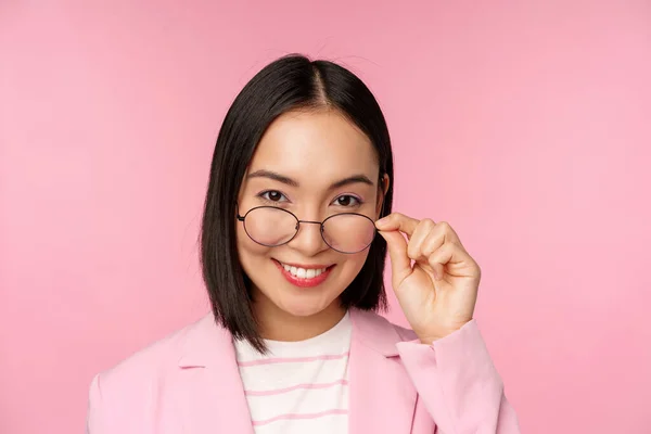 Retrato de mulher de negócios asiática em óculos, olhando intrigado para a câmera e sorrindo, vendedora profissional olhando com interesse, fundo rosa — Fotografia de Stock