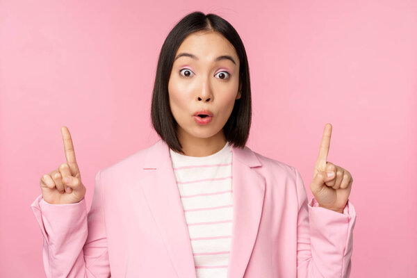 Portrait of asian businesswoman, corporate worker in suit, looks surprised by advertisement, points finger up, showing banner or logo on top, stands over pink background