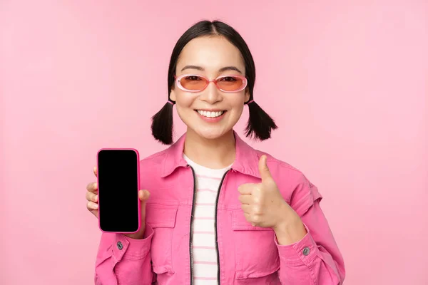 Glad snygg tjej rekommenderar ansökan på mobiltelefon. Leende asiatisk kvinnlig modell visar smartphone skärm och tumme upp, står över rosa bakgrund — Stockfoto