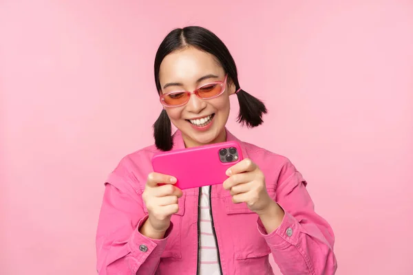 Портрет счастливой азиатской девушки, играющей на смартфоне, смотрящей видео на мобильном телефоне, стоящей на розовом фоне — стоковое фото