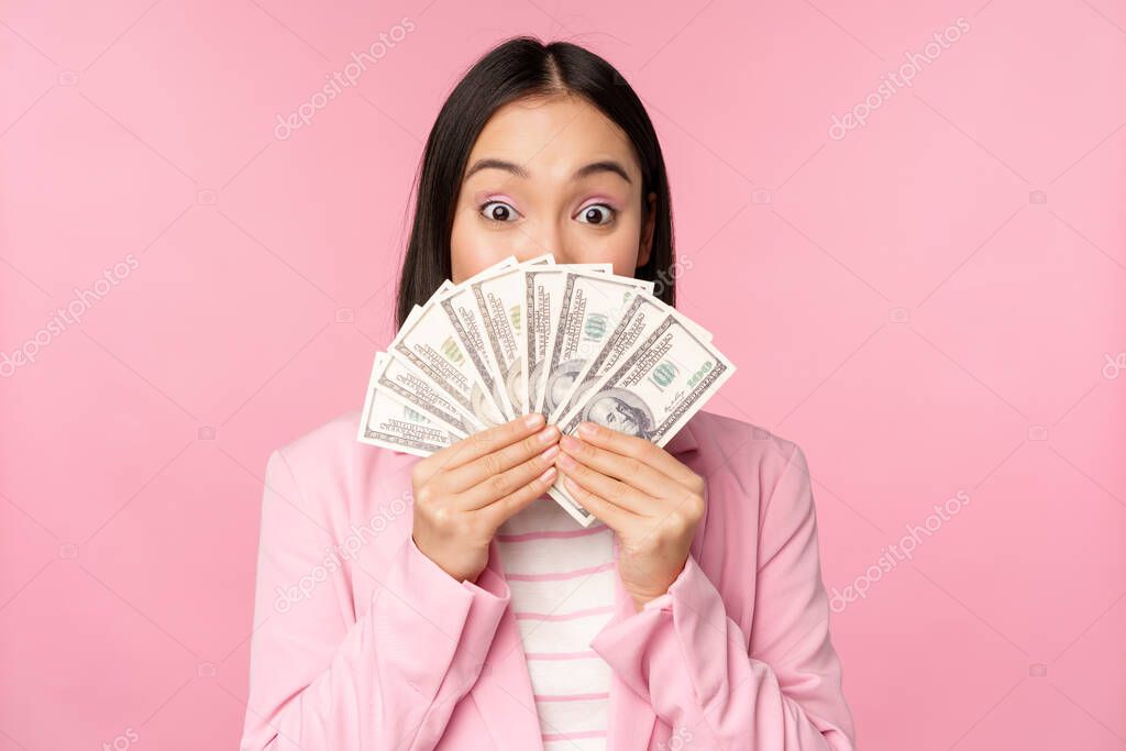 Happy asian lady in suit holding money, dollars with pleased face expression, standing over pink background. Copy space