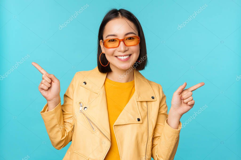 Happy stylish chinese girl in sunglasses, points fingers sideways, invites to check out, see store discount, shows left and right, stands over blue background.