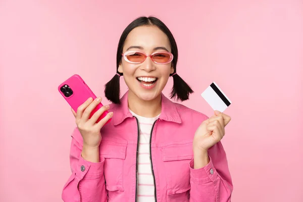 Moderna bella ragazza asiatica, ridendo e sorridendo con il telefono cellulare, carta di credito, shopping online, pagare con smartphone, in piedi su sfondo rosa — Foto Stock