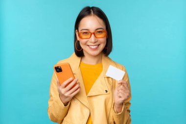 İnternetten alışveriş. Güneş gözlüklü şık genç Asyalı kadın, kredi kartı gösteriyor ve akıllı telefon kullanıyor, internetten ödeme yapıyor, satın alıyor, mavi arka planda duruyor.