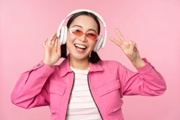 Taniec stylowa azjatycka dziewczyna słuchanie muzyki w słuchawkach, pozowanie na różowym tle — Zdjęcie stockowe