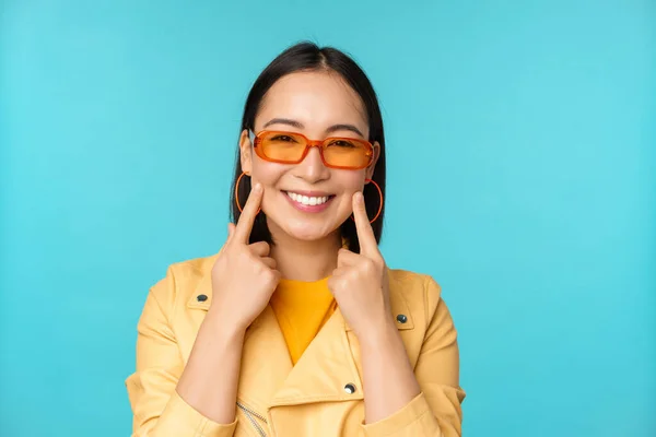 Güneş gözlüklü, gülümseyen ve romantik görünen Asyalı genç bir kadının mavi arka planda mutlu mesut duran portresi. — Stok fotoğraf