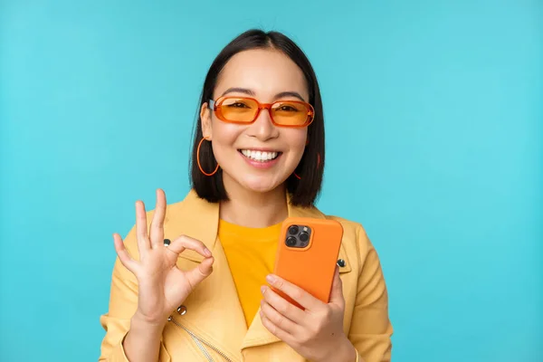 Szczęśliwa koreańska dziewczyna w okularach przeciwsłonecznych, pokazująca w porządku znak i trzymająca telefon komórkowy, korzystająca z aplikacji na smartfona, polecająca aplikację, stojąca nad niebieskim tłem — Zdjęcie stockowe