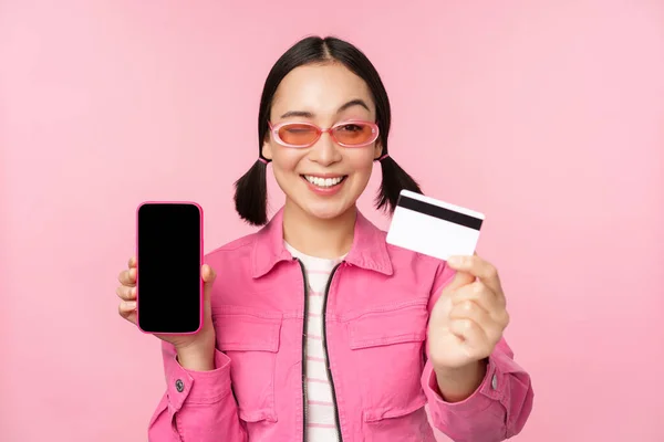 Imagen de una mujer coreana sonriente que muestra la tarjeta de crédito y la pantalla del teléfono móvil, interfaz de la aplicación del teléfono inteligente, pago en línea, compras sin contacto, de pie sobre el fondo rosa — Foto de Stock