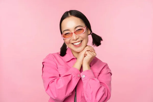 Close up retrato de mulher asiática elegante em óculos de sol, sorrindo, olhando lisonjeado, rindo coquete, de pé sobre fundo rosa — Fotografia de Stock
