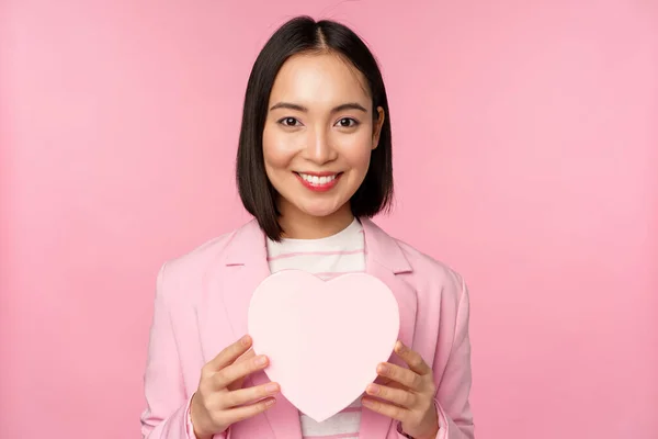 Αυτό είναι για σένα. Ρομαντικό χαριτωμένο ασιατική εταιρική γυναίκα, κορίτσι με κοστούμι, δείχνει καρδιά σε σχήμα κουτί με δώρο, στέκεται πάνω από ροζ φόντο — Φωτογραφία Αρχείου