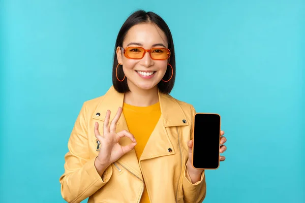 Leende koreansk kvinna visar mobiltelefon app gränssnitt, smartphone ansökan, rekommendera på mobiltelefon, står över blå bakgrund — Stockfoto
