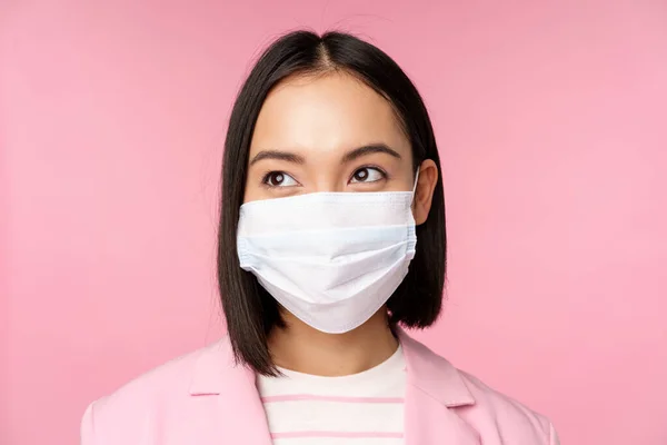 Close up portrait of japanese corporate woman in medical face mask from covid-19, looking left at logo, sale promo, standing over pink background — Stock Photo, Image