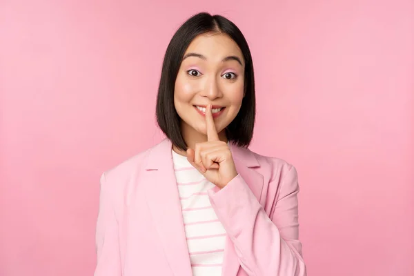Silencio, concepto tabú. Retrato de una mujer de negocios asiática mostrando un gesto de silencio, un signo de shhh, presiona el dedo contra los labios, de pie sobre un fondo rosa en traje —  Fotos de Stock
