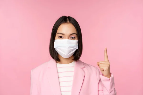 Close up portrait of asian businesswoman in medical face mask and suit, pointing finger up, showing advertisement, top banner, standing over pink background — Stock Photo, Image