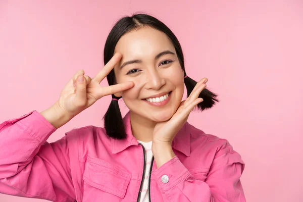 Κοντινό πλάνο της κομψής Ασιάτισσας χαμογελά ευτυχισμένη, δείχνει την ειρήνη v-σημάδι, Kawaii θέτουν, ποζάρουν σε ροζ φόντο — Φωτογραφία Αρχείου