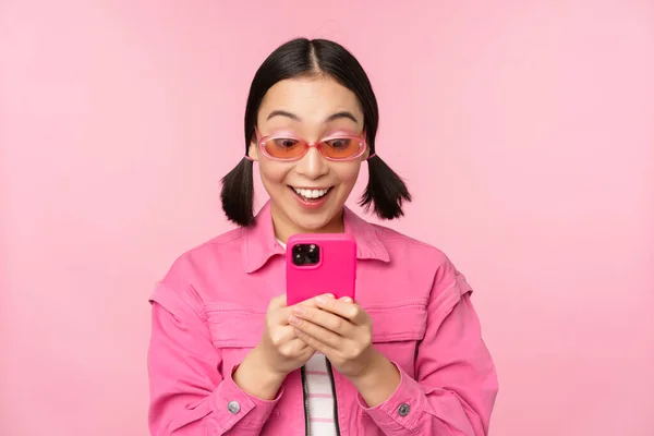 Πορτρέτο της ασιάτισσας με γυαλιά ηλίου χρησιμοποιώντας smartphone. Γυναίκα κοιτάζοντας το κινητό τηλέφωνο, περιήγηση στην εφαρμογή, στέκεται πάνω από ροζ φόντο — Φωτογραφία Αρχείου