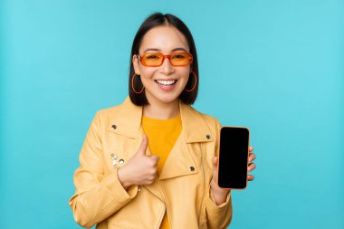 Genç, şık, Çinli bir kadın cep telefonu ekranı ve başparmağını kaldırıyor. Akıllı telefon uygulaması, mavi arka planda duruyor.