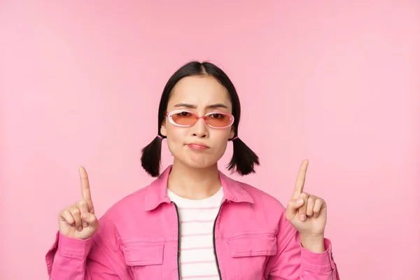 Detailní záběr stylové korejské dívky, vypadá zmateně, ukazuje prsty nahoru, ukazuje reklamy, stojí nad růžovým pozadím — Stock fotografie
