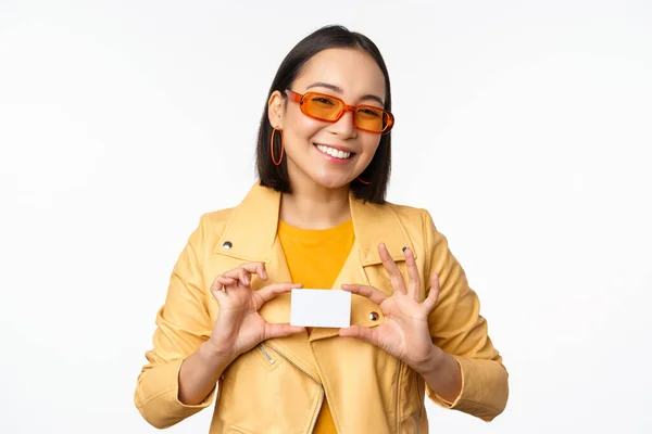 Elegante chica asiática atractiva en gafas de sol, mostrando la tarjeta de crédito y sonriendo, de pie feliz contra fondo blanco estudio — Foto de Stock