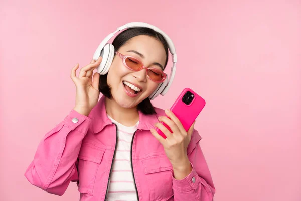 スタイリッシュなアジアの女の子は、スマートフォンで踊ります,携帯電話のアプリでヘッドフォンで音楽を聞きます,笑顔と笑い,ピンクの背景にポーズ — ストック写真