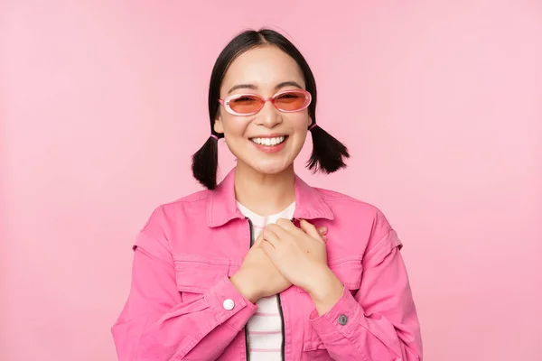 Sorrindo jovem mulher asiática em óculos de sol olhando grato, de mãos dadas no coração, lisonjeado, apreciar smth, de pé sobre fundo rosa — Fotografia de Stock