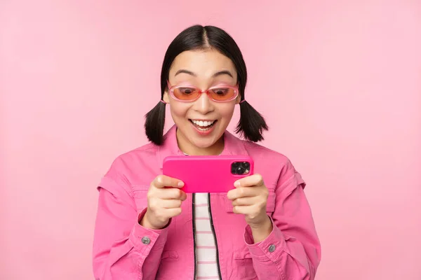 Porträtt av glad asiatisk flicka som spelar på smartphone, titta på videor på mobiltelefon app, står över rosa bakgrund — Stockfoto
