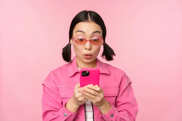 Portret Azjatki w okularach przeciwsłonecznych za pomocą smartfona. Kobieta patrząca na telefon komórkowy, przeglądająca aplikację, stojąca nad różowym tłem — Zdjęcie stockowe