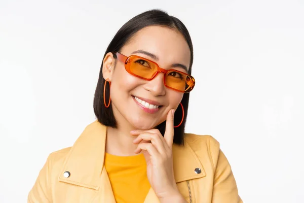 Close up retrato de mulher asiática pensando, vestindo óculos de sol e sorrindo, olhando para cima pensativo, de pé sobre fundo estúdio branco — Fotografia de Stock