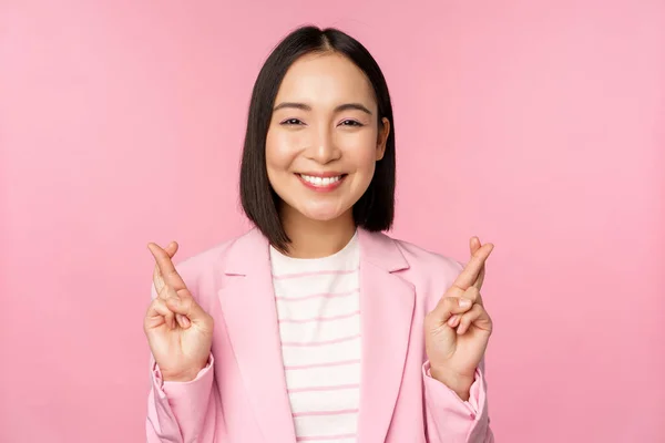 Feliz asiático empresária cruz dedos para boa sorte, desejando, orando e esperando, sorrindo para a câmera, de pé em terno sobre rosa fundo — Fotografia de Stock
