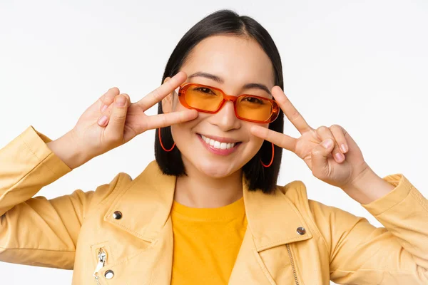 Close up retrato de jovem mulher asiática, menina elegante em óculos de sol, mostrando paz, v-sign e sorrindo, de pé sobre fundo estúdio branco — Fotografia de Stock