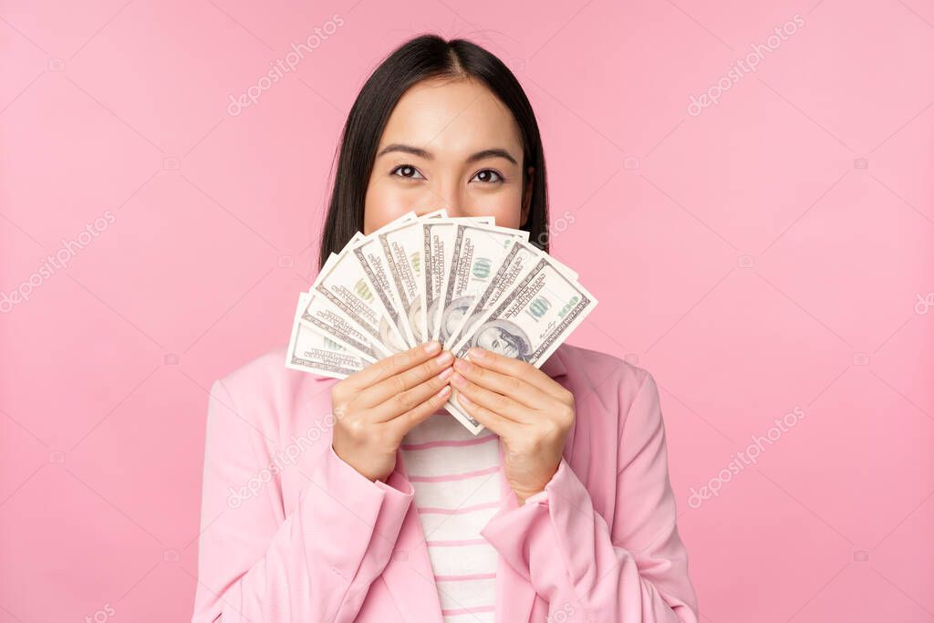 Happy asian lady in suit holding money, dollars with pleased face expression, standing over pink background. Copy space