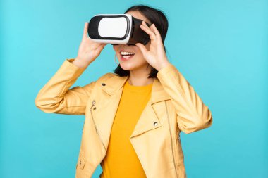 VR kulaklık takan mutlu Asyalı kadın modelin portresi. Sanal gerçeklik gözlükleriyle gülüyor ve gülümsüyor. Mavi arka planda duruyor.