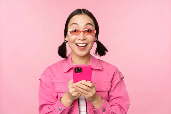 Ritratto di ragazza coreana con eleganti occhiali da sole che reggono il telefono cellulare, utilizzando l'app per smartphone, in piedi su sfondo rosa — Foto Stock