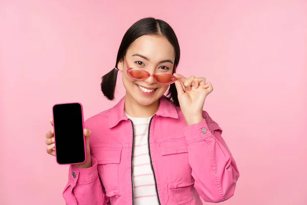 Portrait of stylish modern asian girl shows mobile phone screen, smartphone app interface, standing in sunglasses against pink background — Stock Photo, Image