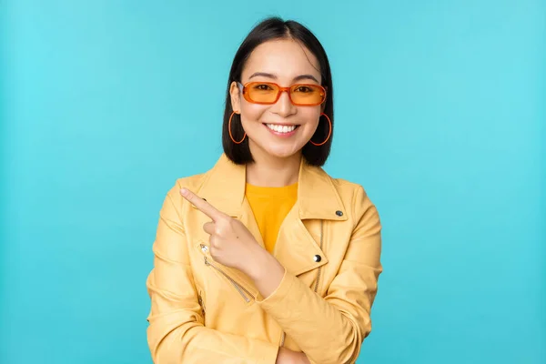Elegante mujer asiática sonriente en gafas de sol, señalando con el dedo a la izquierda, mostrando pancarta publicitaria, de pie en el abrigo amarillo de moda sobre fondo azul — Foto de Stock