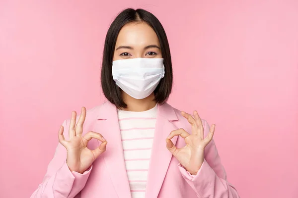 Asian businesswoman in suit and medical face mask, showing okay sign, recommending using protective equipment in office during covid-19 pandemic, pink background — Stock Photo, Image