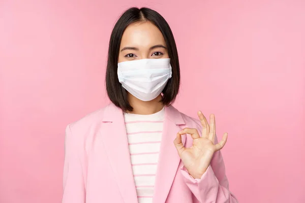 Portrait of asian businesswoman in medical face mask, showing okay sign, wearing suit, work rules during covid-19 pandemic, standing over pink background — Stock Photo, Image