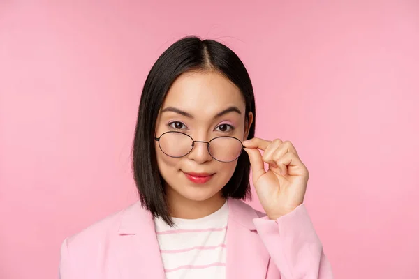 Retrato de mulher de negócios asiática em óculos, olhando intrigado para a câmera e sorrindo, vendedora profissional olhando com interesse, fundo rosa — Fotografia de Stock