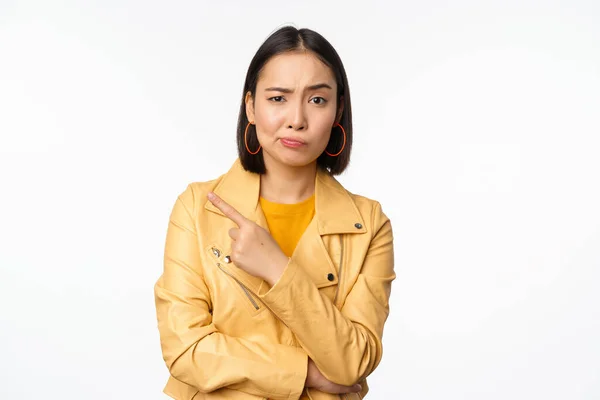 Retrato de elegante menina asiática olhando com dúvida, rosto cético, apontando o dedo esquerdo para o logotipo ou propaganda, de pé contra o fundo branco — Fotografia de Stock