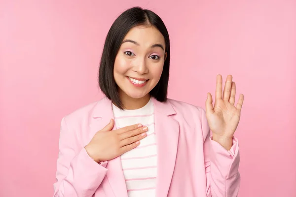 Retrato de bonito asiático empresária levantando mão, apresentar-se no escritório, sorrindo coquete, de pé sobre fundo rosa no terno — Fotografia de Stock