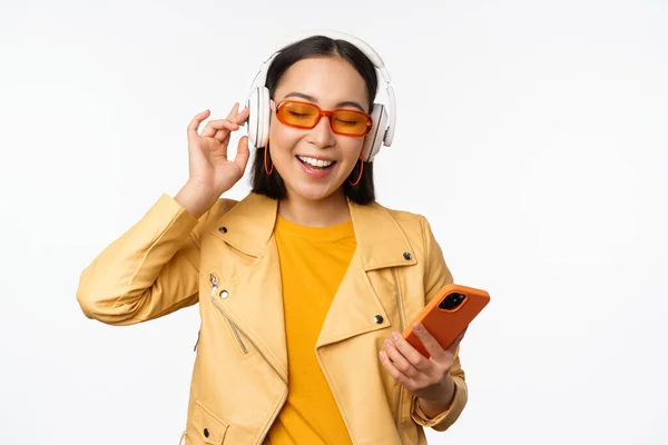 Feliz mujer asiática con estilo en gafas de sol, escuchando música en auriculares, sosteniendo el teléfono inteligente, cantando y bailando, de pie sobre fondo blanco — Foto de Stock