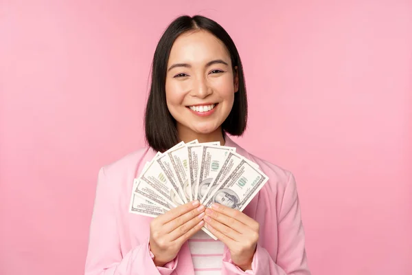 Ekonomi, mikrokrediter och människor koncept. Glad leende asiatisk affärskvinna visar dollar pengar, står i kostym mot rosa bakgrund — Stockfoto