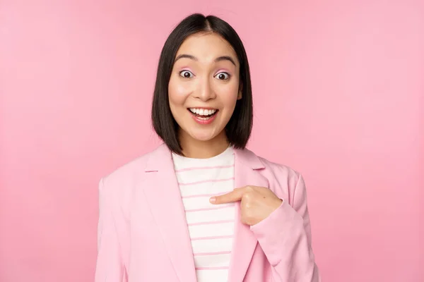 Retrato de jovem mulher de negócios asiática com surpreso, excitado expressão facial, apontando o dedo para si mesma, de pé em terno sobre fundo rosa — Fotografia de Stock