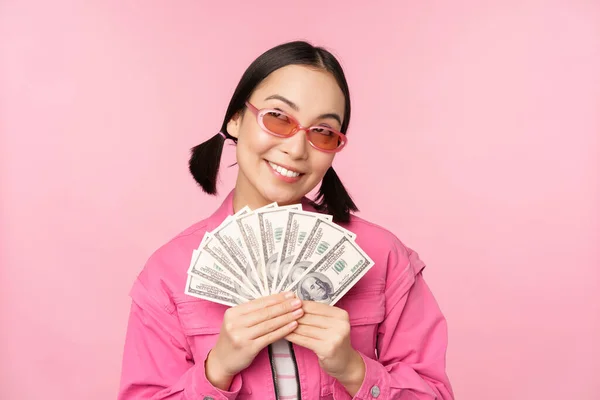 Beautiful korean woman in sunglasses, showing dollars, money cash, smiling pleased, concept of fast loans, microcredit and payment, standing over pink background — Stock Photo, Image