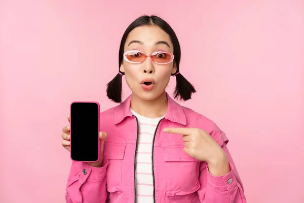 Entuzjastyczna Azjatka w stylowych ubraniach, okularach przeciwsłonecznych, wskazująca palcem na ekran telefonu komórkowego, pokazująca aplikację na smartfona, stojąca nad różowym tłem — Zdjęcie stockowe