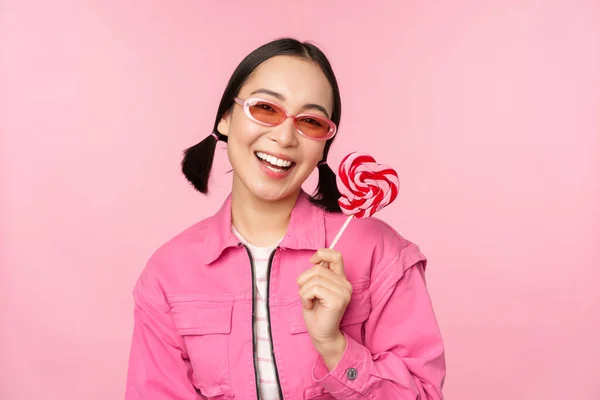 Κομψό κορεάτικο κορίτσι γλείφει γλειφιτζούρι, τρώει γλυκά και χαμογελά, στέκεται με γυαλιά ηλίου σε ροζ φόντο — Φωτογραφία Αρχείου