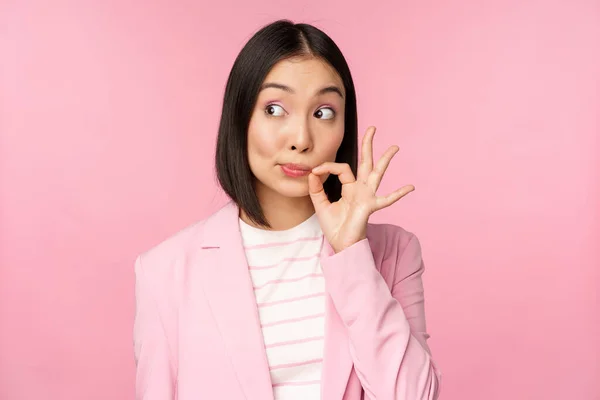 Retrato de mujer corporativa asiática mostrando sello bucal, cerrar los labios en el gesto clave, promesa mantener en secreto, de pie sobre fondo rosa en traje —  Fotos de Stock