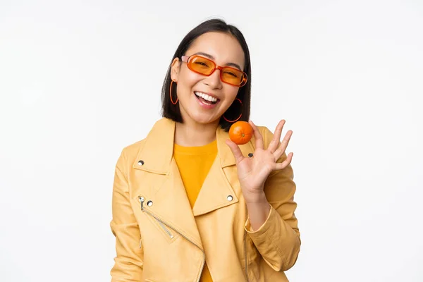 Hermosa chica asiática en gafas de sol mostrando mandarina y sonriente, luciendo feliz, posando en amarillo sobre fondo de estudio — Foto de Stock