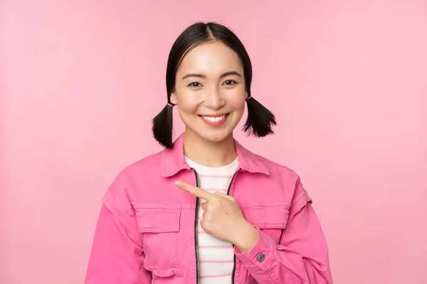 Retrato de bela menina asiática sorridente, apontando o dedo esquerdo, mostrando propaganda, banner promo, de pé sobre fundo rosa — Fotografia de Stock