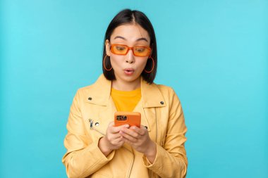 Güneş gözlüklü Asyalı bir kızın portresi, etkilenmiş, şaşkın yüz ifadesi ile akıllı telefon ekranına bakıyor, vay be yüz, mavi arka planda duruyor.